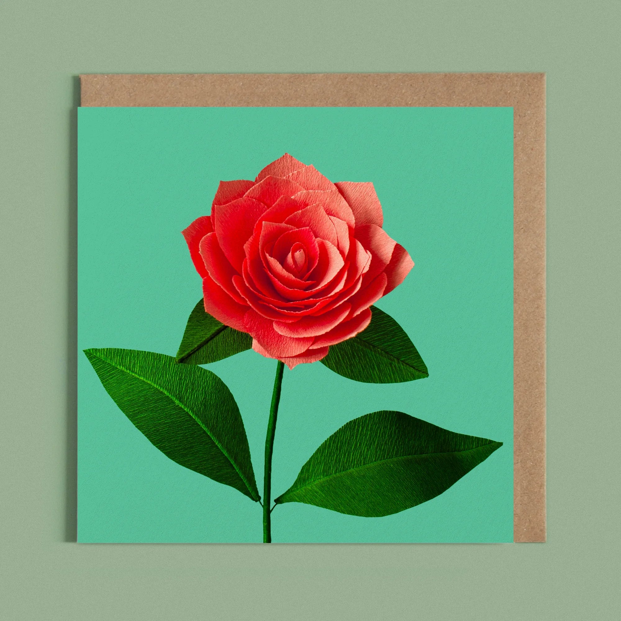 Greeting card with a image of a handcrafted paper coral camellia on a green background by paper artist Daphne Fitch.