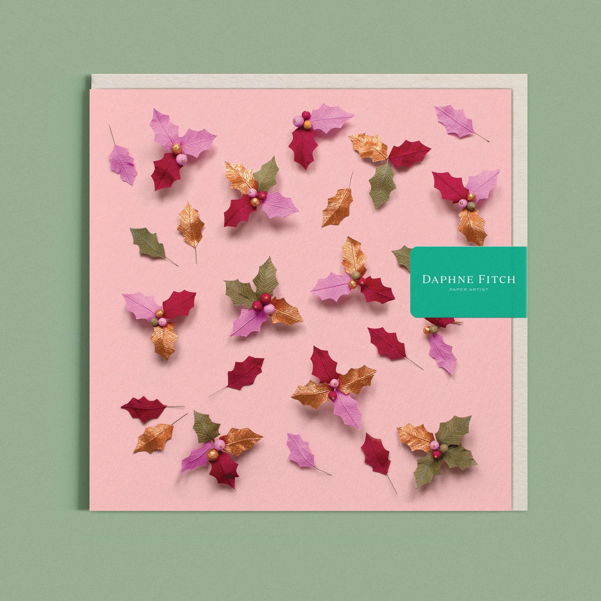 Greeting card with a image of a handcrafted paper holly leaves and berries on a pink background by paper artist Daphne Fitch.